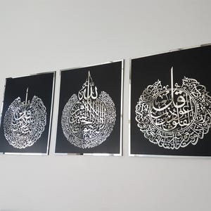 May include: Three framed wall art pieces with intricate silver calligraphy on a black background. The artwork is displayed on a white wall. Each piece has a silver frame, adding a touch of elegance to the Islamic art.
