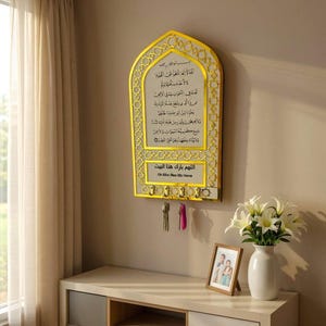 Puede incluir: Un llavero decorativo montado en la pared con un marco dorado y caligrafía árabe. El llavero tiene varios ganchos y un diseño rectangular con patrones ornamentados. El texto "Oh Allah Bless this Home" es visible.