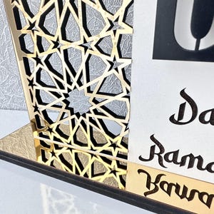 May include: A gold and black wooden Ramadan calendar with an intricate geometric design. The calendar features a white card with the text "Day Ramadan" written in black calligraphy.