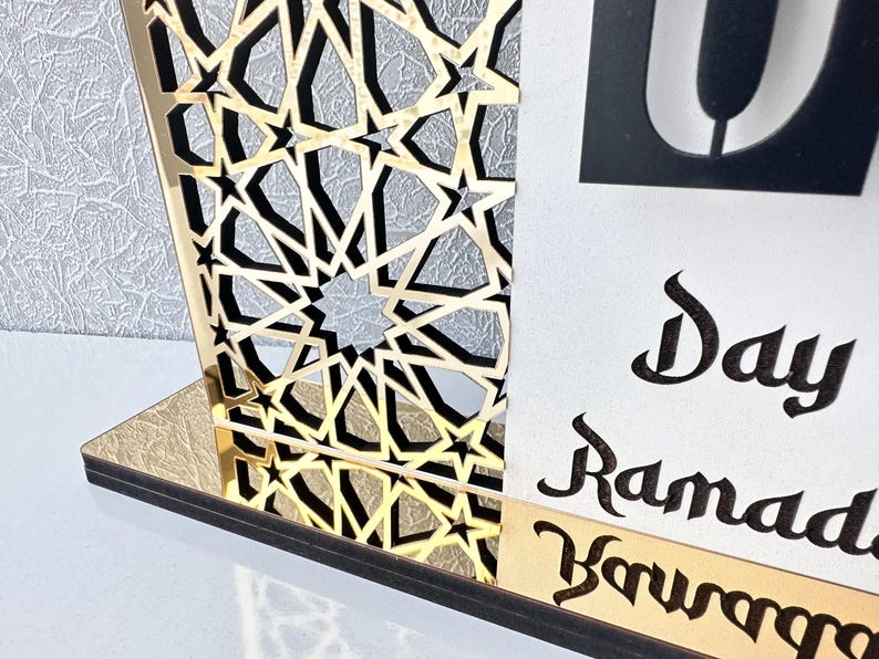 May include: A gold and black wooden Ramadan calendar with an intricate geometric design. The calendar features a white card with the text "Day Ramadan" written in black calligraphy.