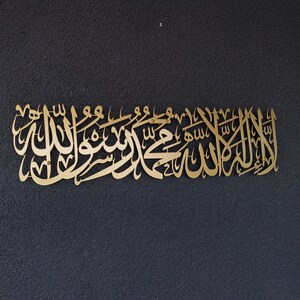 Może przedstawiać: Złote, prostokątne dzieło sztuki ściennej z arabską kaligrafią. Skrypt jest misternie zaprojektowany i zamontowany na ciemnoszarym, teksturowanym tle. Grafika to element dekoracyjny do wystroju wnętrz.