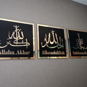 May include: Three gold framed wall plaques with black backgrounds and gold Arabic calligraphy. The plaques read "Allahu Akbar", "Alhamdulillah", and "Subhanallah".
