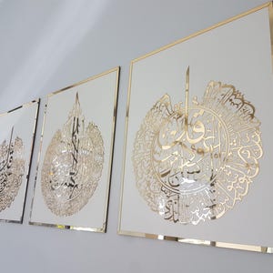 May include: Three framed wall art pieces with gold-colored calligraphy on a white background. The frames have a gold border. The artwork is displayed on a light gray wall.