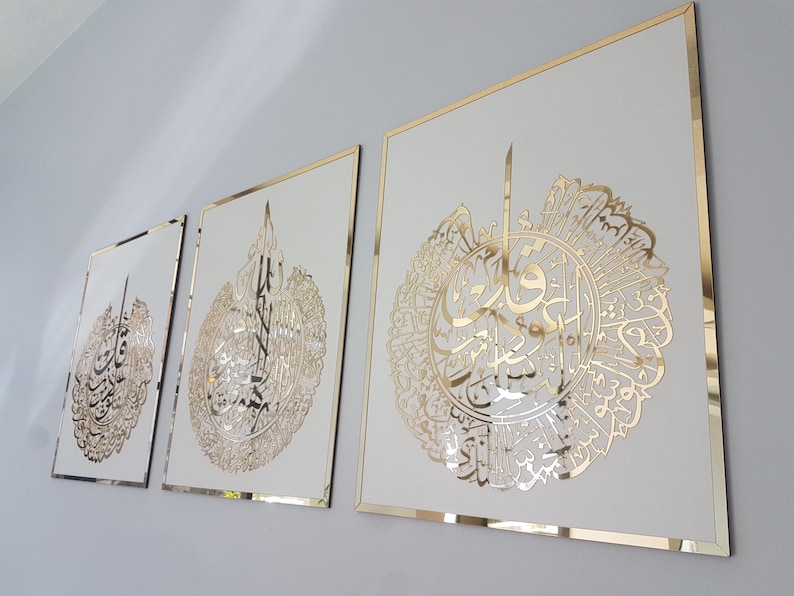 May include: Three framed wall art pieces with gold-colored calligraphy on a white background. The frames have a gold border. The artwork is displayed on a light gray wall.