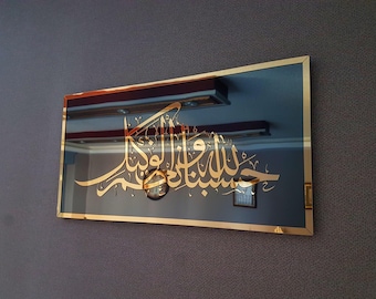 May include: A rectangular mirror with a gold frame and gold Arabic calligraphy. The mirror reflects the room's interior, including lights and a clock. The mirror is mounted on a textured gray wall.