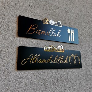 May include: Two black rectangular signs with gold lettering. The top sign says "Start with Bismillah" with a fork and spoon icon. The bottom sign says "End with Alhamdulillah" with two hands in a praying position.