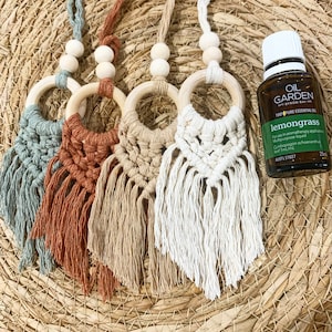 May include: Four macrame car air fresheners with wooden rings and beads, hanging from a string. The air fresheners are in different colours: teal, orange, beige, and white. A bottle of lemongrass essential oil is in the background.