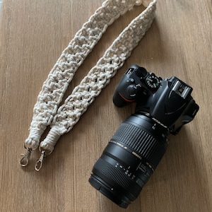 Puede incluir: Una correa de cámara de crochet blanco con cierres plateados y una cámara DSLR negra con un objetivo.
