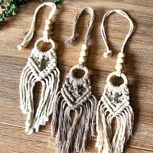 May include: Three macrame wall hangings with wooden beads and rings. The hangings are made with natural cotton cord and feature different coloured tassels. Each hanging has a small metal charm in the centre.