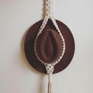 May include: A brown felt hat suspended by a cream-coloured macrame cord hanger. The hanger features a circular wooden ring at the top for mounting and a decorative knotted pattern that cradles the hat, finishing with a long fringe tassel at the bottom.
