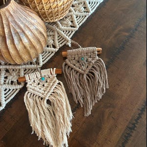 Puede incluir: Dos pequeñas tapices de macramé de pared con listones de madera y detalles turquesa. Los tapices están hechos con hilo de color natural y tienen detalles de flecos.