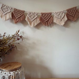 May include: A macrame banner with beige and brown triangular flags hanging on a white wall. The banner is made of cotton cord and has a rustic, bohemian style.