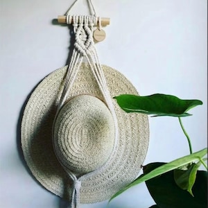 May include: A cream-coloured macrame hat hanger featuring a wooden dowel and a small circular wooden tag. The hanger is constructed from braided cotton cord and supports a wide-brimmed straw hat with a woven texture, suspended against a wall.
