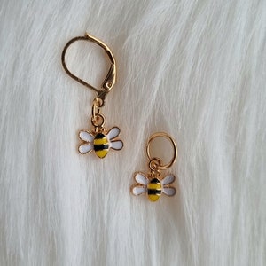 Puede incluir: Dos marcadores de punto de abeja de oro. Los abalorios de abeja son blancos con rayas negras y cuerpos amarillos. Los marcadores de punto están sobre un fondo blanco peludo.