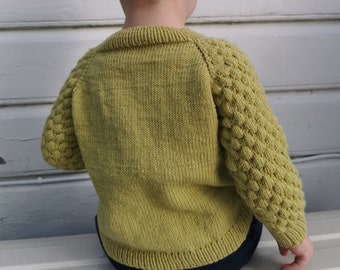 Può includere: Un bambino indossa un maglione di lana verde con maniche a trama. Il maglione ha un collo rotondo e maniche lunghe.