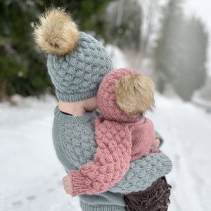 Könnte beinhalten: Eine Person trägt eine hellblaue Strickmütze und einen Pullover und hält ein Kind in einer rosa Strickmütze, einem Schal und einem Pullover. Beide Mützen haben einen beigefarbenen Kunstfell-Bommel. Verschneite Winterlandschaft.