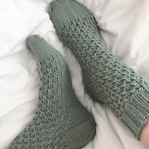 Könnte beinhalten: Ein Paar salbeigrüne Stricksocken mit einem strukturierten Muster. Die Socken liegen auf einem weißen Bettlaken.