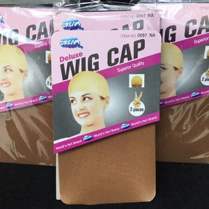Può includere: Più confezioni di cuffie per parrucche Deluxe. Ogni confezione presenta un'etichetta rosa e bianca con la parola "DREAM" e il testo "Deluxe Wig Cap". Le confezioni contengono due pezzi ciascuna. Le cuffie per parrucche sono di colore marrone chiaro.