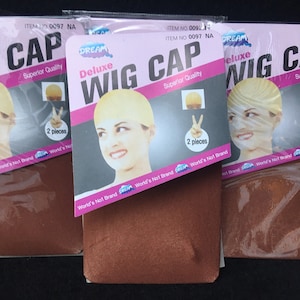 Può includere: Sono esposte più confezioni di cuffie per parrucche Deluxe. Ogni confezione presenta un'etichetta rosa e bianca con la scritta "Deluxe Wig Cap" e "Superior Quality". Le confezioni contengono due cuffie per parrucca ciascuna. Lo sfondo è nero.