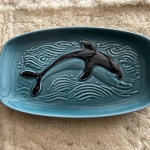 Könnte beinhalten: Eine rechteckige, türkisfarbene Keramikschale mit einem erhabenen, schwarzen Delfin-Design. Die Schale weist ein Wellenmuster um den Delfin herum auf, was ein nautisches Thema hinzufügt. Der Delfin befindet sich in einer Sprungpose.
