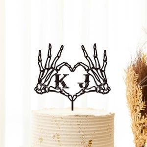 May include: Black skeleton hands forming a heart shape with the initials "K" and "J" on a white cake topper. The topper is on a white cake with gold accents.