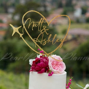 Puede incluir: Una tarta de boda blanca de varios pisos adornada con flores rosas y burdeos. Una decoración para tarta de corazón con purpurina dorada con los nombres "Matt & Ashley" está colocada encima. Fondo verde borroso.