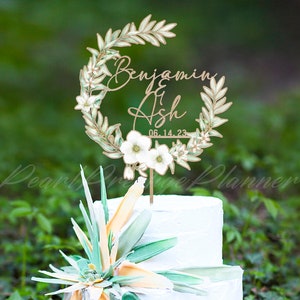 Peut inclure: Un gâteau de mariage blanc à deux niveaux avec un décor de gâteau. Le décor est une couronne en bois avec les noms "Benjamin & Ash" et la date "06.14.23". Le gâteau est décoré d'un grand arrangement floral coloré.