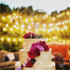 May include: A rose gold cake topper shaped like a heart with an airplane silhouette on the left side. The text "Sophia is 16" is written in a cursive script inside the heart. The topper is placed on a three-tiered cake decorated with red roses.