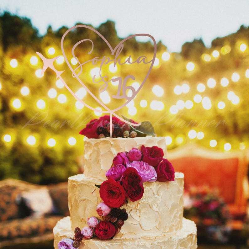 May include: A rose gold cake topper shaped like a heart with an airplane silhouette on the left side. The text "Sophia is 16" is written in a cursive script inside the heart. The topper is placed on a three-tiered cake decorated with red roses.