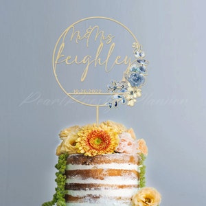 May include: A gold-colored cake topper with the words "Mrs Keighley" and the date "10.26.2022" in a circular design. The design includes blue floral accents. The topper is placed on a cake decorated with yellow and orange flowers.