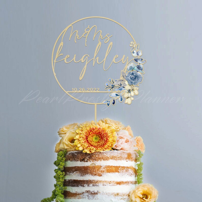 May include: A gold-colored cake topper with the words "Mrs Keighley" and the date "10.26.2022" in a circular design. The design includes blue floral accents. The topper is placed on a cake decorated with yellow and orange flowers.