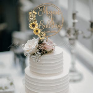 Puede incluir: Una tarta de boda blanca de dos pisos coronada con un adorno de pastel de madera. El adorno presenta los nombres "Megan & Sabrina" y la fecha "11.09.2023" dentro de un dise&ntilde;o circular, adornado con girasoles y detalles florales.
