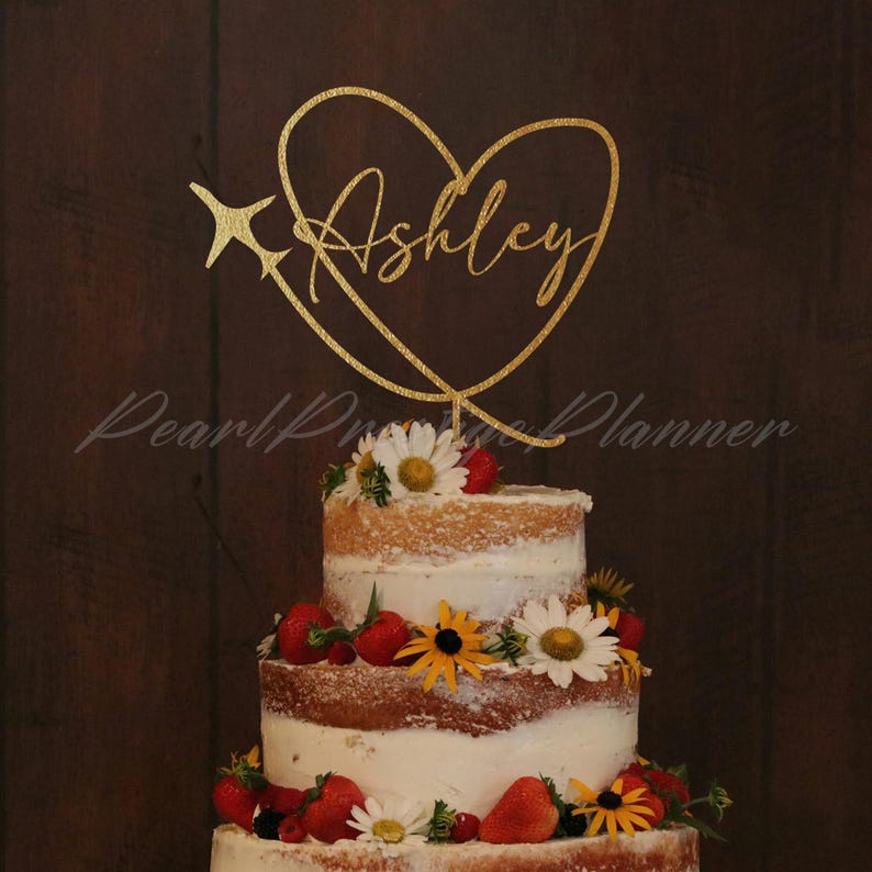 May include: A gold-colored cake topper featuring a heart outline, a small airplane silhouette, and the name Ashley in cursive script, positioned atop a three-tiered naked cake decorated with white daisies and red strawberries.