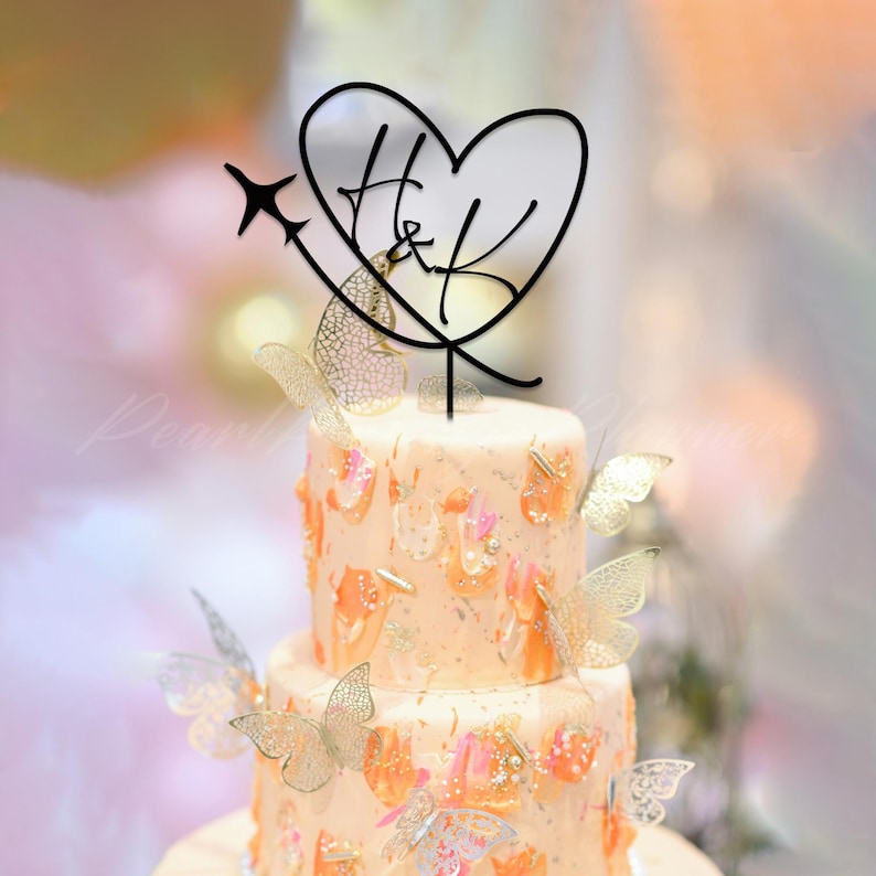 May include: A two-tiered cake decorated with orange and pink brushstrokes, gold glitter, and translucent butterfly cutouts. A black cake topper features a heart outline, the letters H and K, and a small airplane silhouette on a stem.