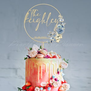 May include: A wedding cake topped with a gold-colored cake topper. The topper features the name "The Keighley" and the date "10.26.2022" within a circular design with floral accents. The cake is decorated with pink and white frosting, strawberries, and macarons.