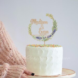 May include: A white frosted cake topped with colorful sprinkles and a wooden cake topper. The topper features the names "Madison & Gann" with a floral wreath design and the date 10.06.2023.