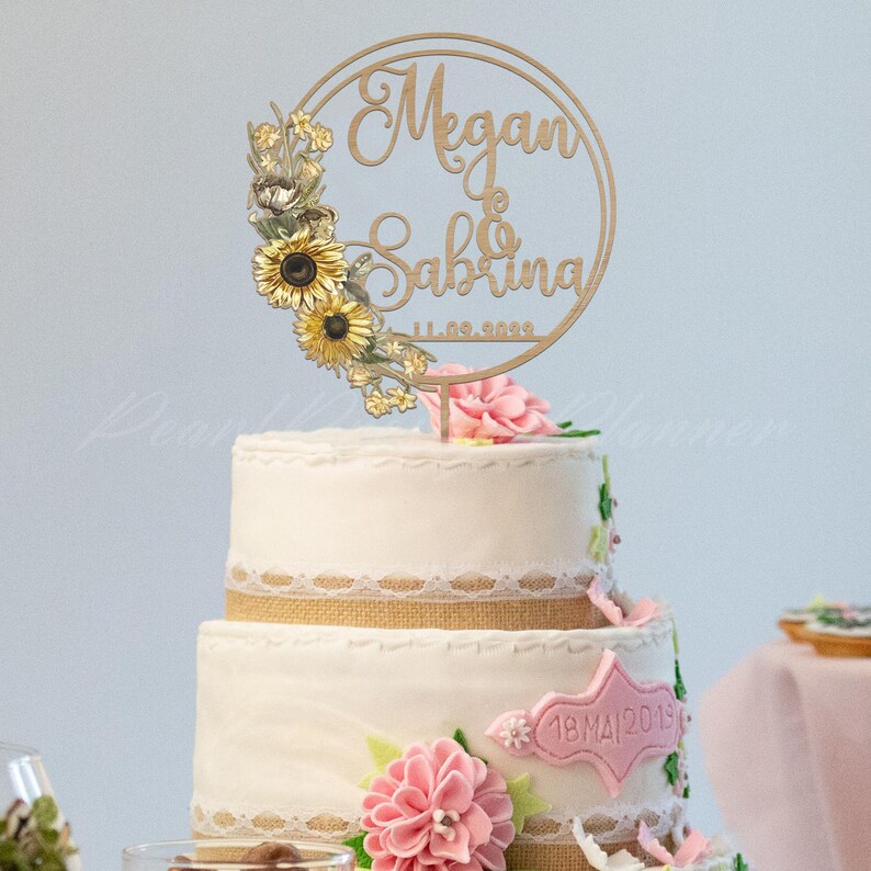 Puede incluir: Una tarta de boda de dos pisos con una decoraci&oacute;n de madera. La decoraci&oacute;n presenta los nombres "Megan & Sabrina" y la fecha "11.09.2020" dentro de un marco circular, adornado con girasoles. La tarta est&aacute; decorada con flores rosas y encaje.
