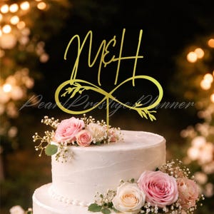 Może przedstawiać: Złoty topper na tort z inicjałami "M&H" w ozdobnym kroju pisma, połączony ze znakiem nieskończoności. Biały, wielopoziomowy tort jest ozdobiony różowymi i kremowymi różami oraz małymi białymi kwiatami. Widoczny jest również tekst "Dear Prestige Planner".