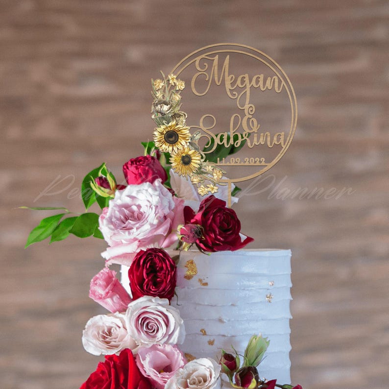 Puede incluir: Una tarta de boda coronada con un adorno circular dorado. El adorno presenta los nombres "Megan & Sabrina" y la fecha "11.09.2022". La tarta est&aacute; decorada con rosas rojas, rosas y blancas, girasoles y detalles de pan de oro.