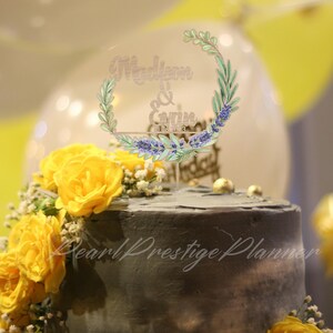 May include: A decorated cake with yellow roses and a clear acrylic topper. The topper features the names "Madison & Gavin" and the date "01.12.23" within a floral wreath design. The cake has a dark gray frosting.