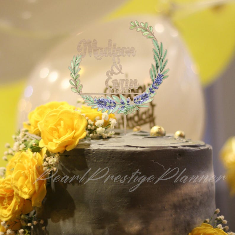 May include: A decorated cake with yellow roses and a clear acrylic topper. The topper features the names "Madison & Gavin" and the date "01.12.23" within a floral wreath design. The cake has a dark gray frosting.