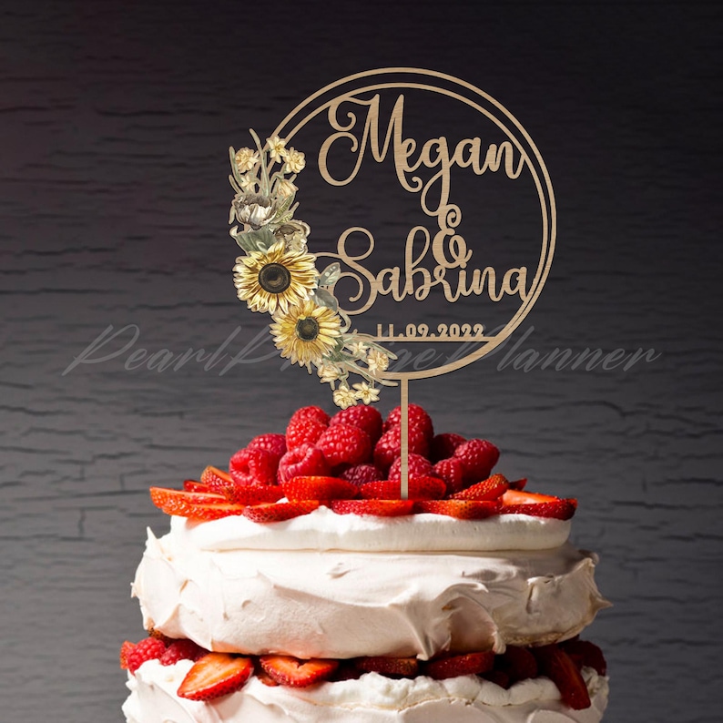 Puede incluir: Un adorno para pastel de madera con los nombres "Megan & Sabrina" y la fecha "11.09.2022" en un dise&ntilde;o circular. El adorno est&aacute; decorado con girasoles y elementos florales. El pastel est&aacute; decorado con fresas y frambuesas.