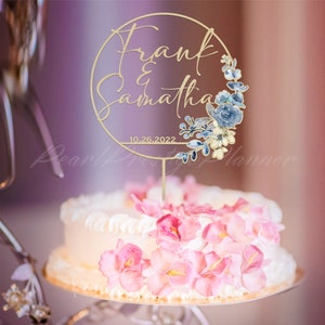 May include: A gold-colored cake topper with the names "Frank & Samantha" and the date "10.26.2022" is displayed on a white cake decorated with pink flowers. The topper has a circular design with floral accents in blue and cream.