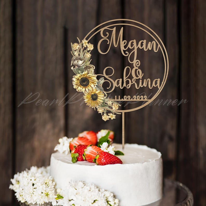 Puede incluir: Un adorno de pastel de madera con los nombres "Megan & Sabrina" y la fecha "11.09.2022" en un dise&ntilde;o circular, adornado con girasoles y vegetaci&oacute;n. El pastel es blanco, cubierto con fresas y flores blancas.