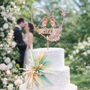 May include: A white tiered wedding cake with a decorative floral arrangement. A heart-shaped cake topper features two birds, floral accents, and the initials "H&K". The background shows a couple kissing, with a blurred floral backdrop.