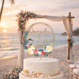 May include: A wedding cake topper with a gold circular frame, featuring the names "Amanda & Clare" and the date "11.05.24". The topper is decorated with watercolor flowers, seashells, and a starfish. The cake is on a beach with a floral arch in the background.