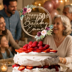 May include: A circular wooden cake topper featuring the text Happy 50th Birthday Emma, decorated with watercolor-style floral accents in shades of deep purple, pink, and green, placed atop a layered cake covered in white cream and fresh raspberries.