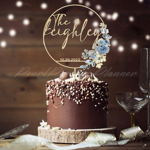 May include: A chocolate cake with white and brown sprinkles, topped with a gold-colored cake topper. The topper features the words "The Keighley" in script, a floral design, and the date "10.26.2022".