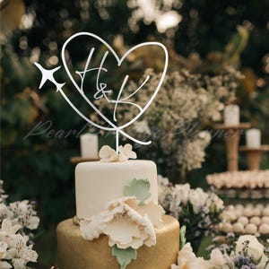May include: A silver-colored cake topper featuring a heart outline with the letters H and K inside, accented by a small airplane silhouette on the left side. The topper is mounted on a thin vertical stem and is positioned atop a two-tiered cake.