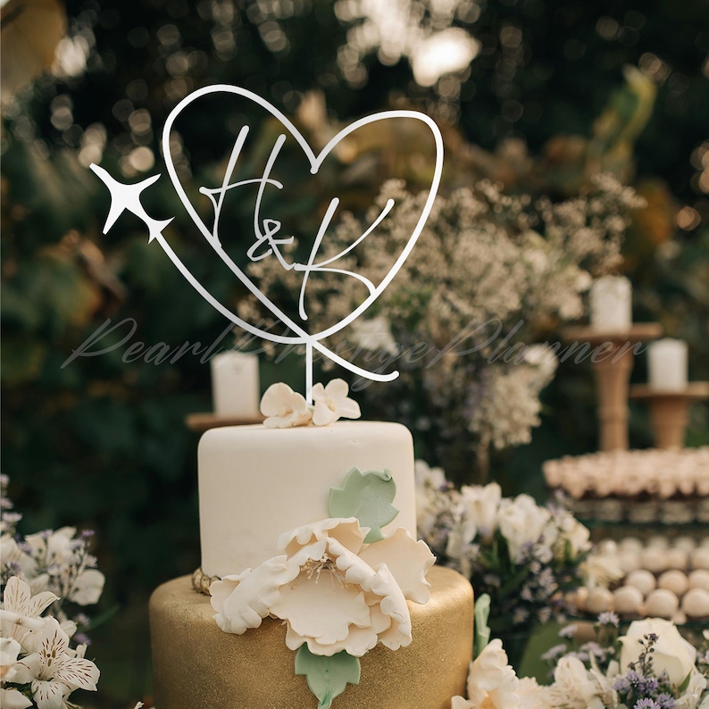 May include: A silver-colored cake topper featuring a heart outline with the letters H and K inside, accented by a small airplane silhouette on the left side. The topper is mounted on a thin vertical stem and is positioned atop a two-tiered cake.
